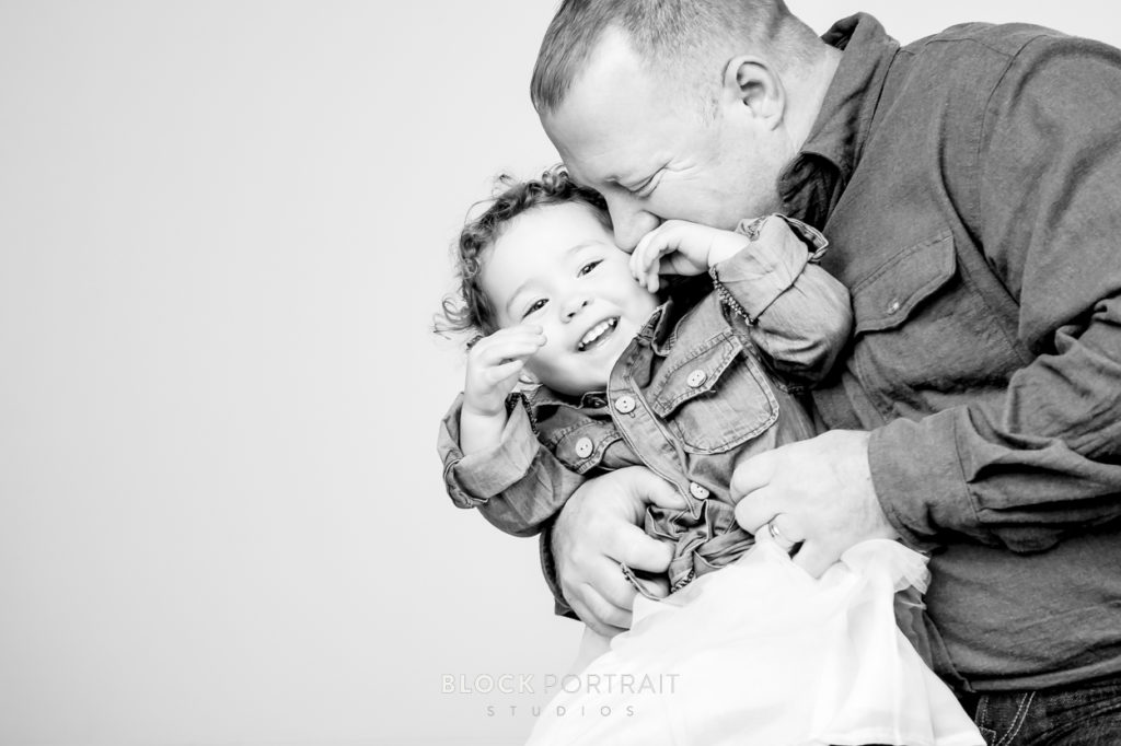 Black and white portrait of a father kissing his daughter on his cheek while she is smiling taken by Block Portrait Studios in Saint Paul Minnesota.
