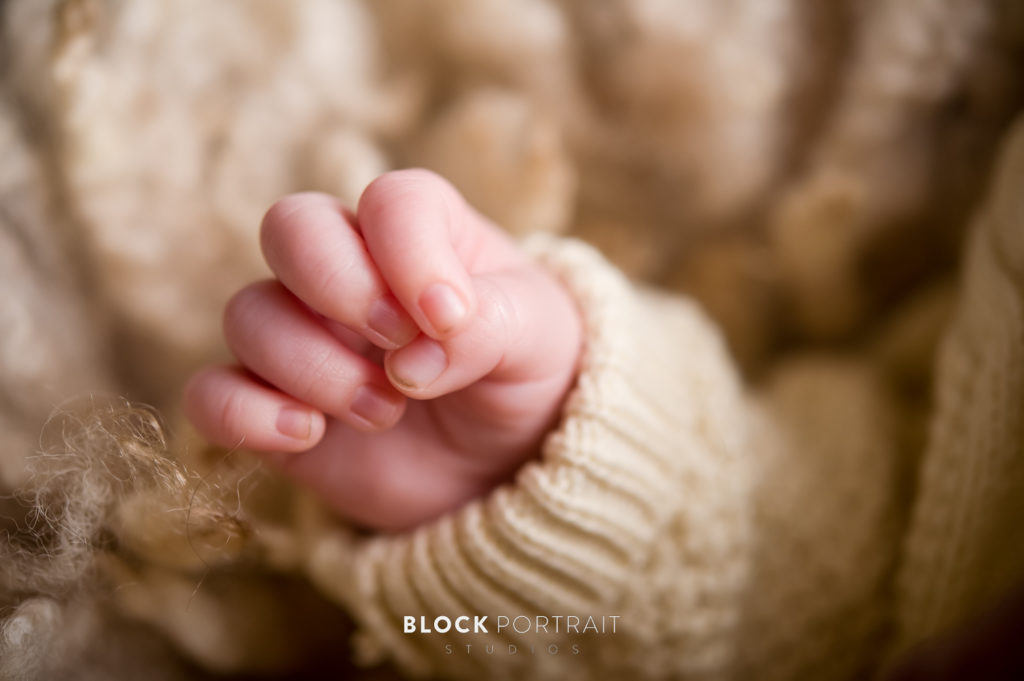 A newborn photograph of a newborn's hand curled in a fist wearing a tan colored sweater taken by newborn photographer Block Portrait Studios.