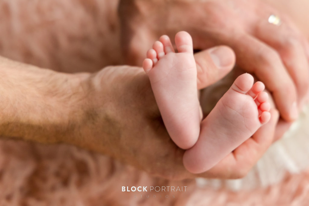 Photo of a newborn's feet and their mother or father's hand caressing them, with tan decor surrounding and accenting the image by Block Portrait Studios.