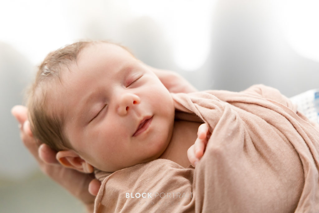 Newborn photograph of newborn baby being held and wrapped in a blanket, with their eyes closed and smiling by St. Paul Photographer Block Portrait Studios.