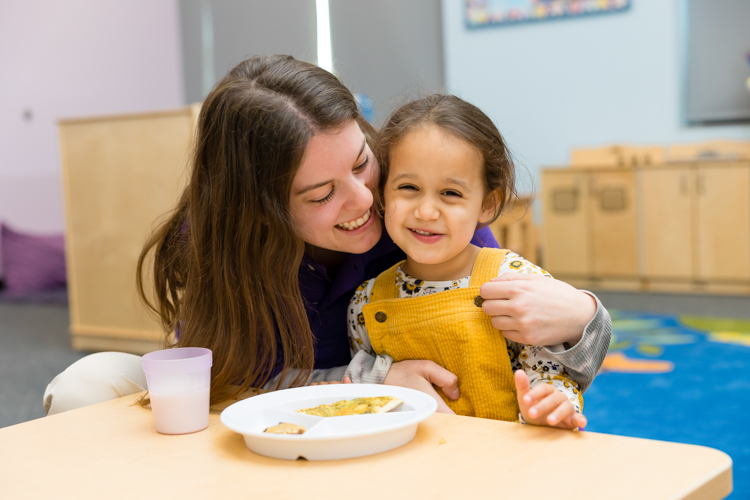 Teacher helping preschool child with lunch and smiling at photographer Block Portrait Studios