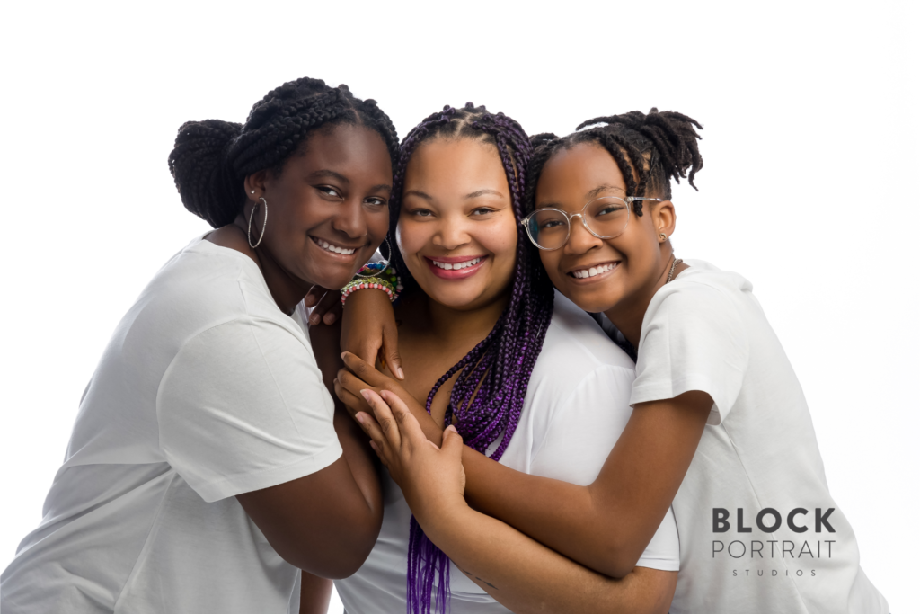 African American mother hugging her two African American teenage daughters, smiling, wearing white t-shirts and standing in a white room.