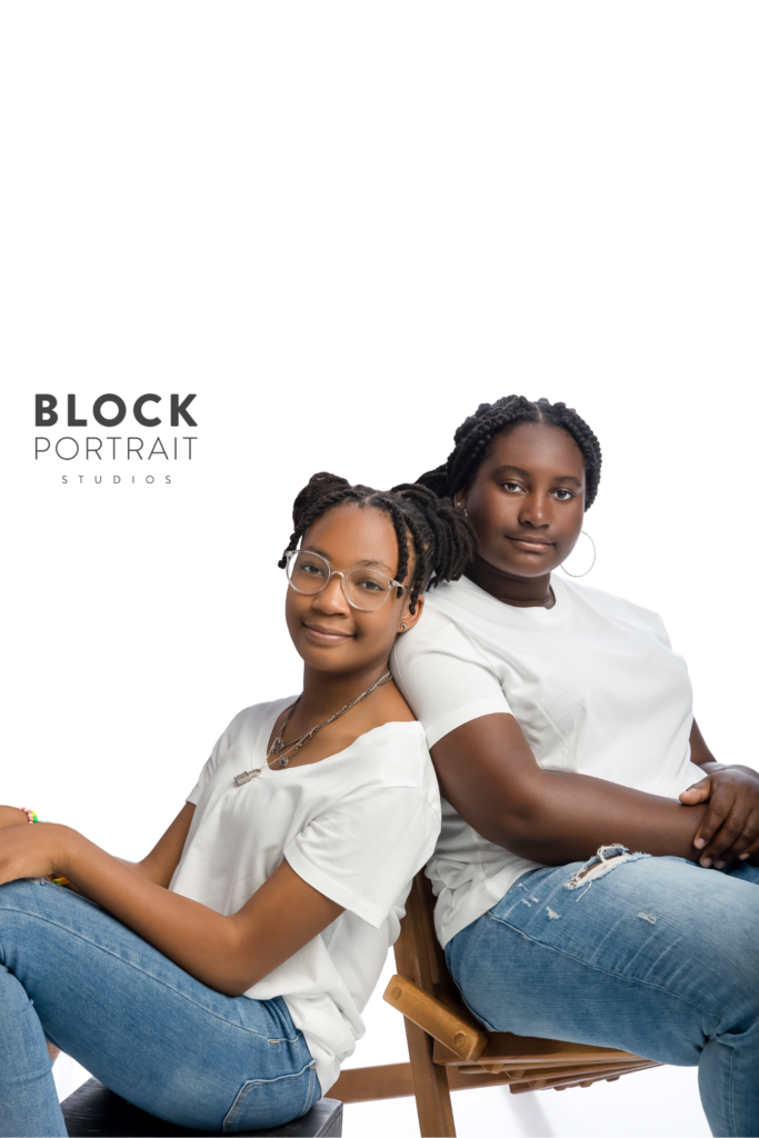 African American teenage sisters, sitting against each other in a white room wearing white t-shirts and denim jeans in an article about helping children after divorce.