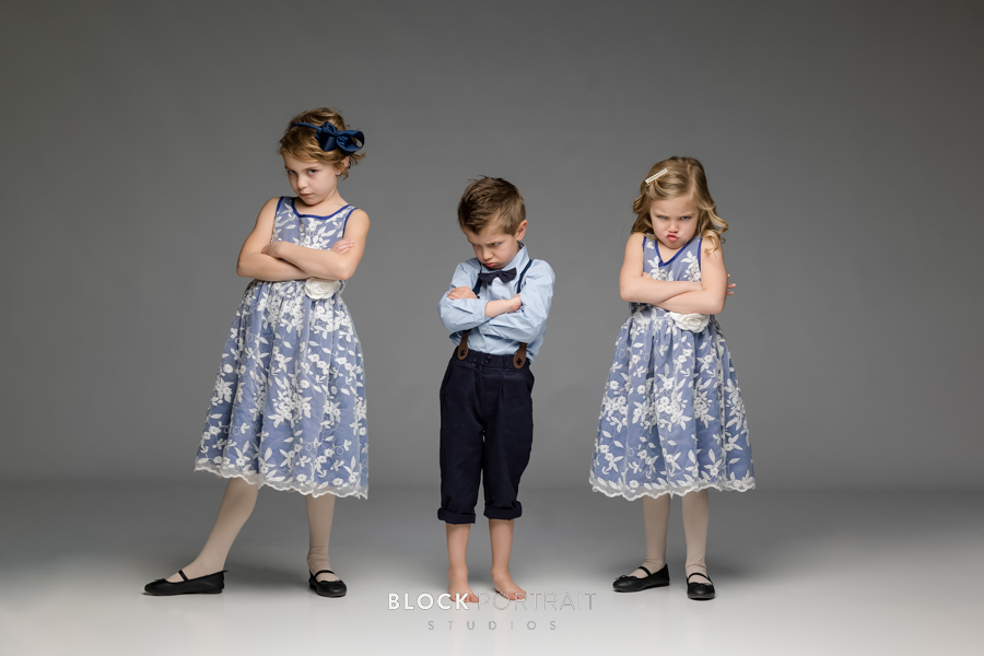 Family portrait of three young siblings, two girls wearing blue dresses and a boy wearing a blue outfit with suspenders and a bow tie, all with pouting facial expressions taking by family portrait photographer.
