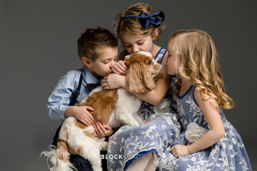 Group of 3 siblings, two girls and a boy, all wearing blue formal attire, holding their pet dog, and giving kisses, by family portrait photographer in St. Paul.