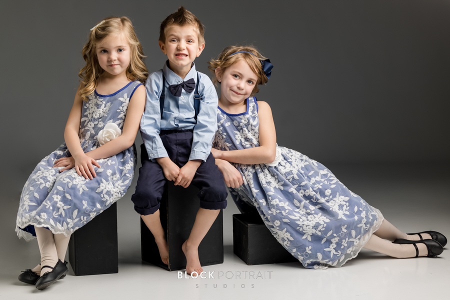 Three young siblings, two girls and a boy, wearing blue dresses and blu e suit, sitting together smiling taken by family portrait photographer.
