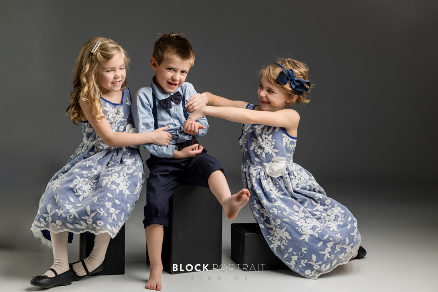 Three siblings, two girls wearing blue floral dresses, and one boy wearing black suspenders and trousers with blue formal shirt, tickling and laughin with each other taking by Minnesota family portrait photographer.