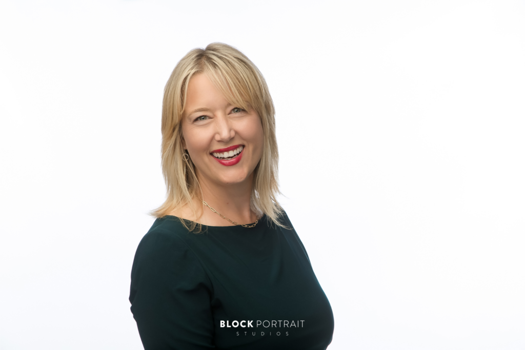 Portrait of a blonde, Caucasian woman, wearing a black, boat neck shirt, smiling at the camera, standing in front of a white background by Twin Cities photographer.