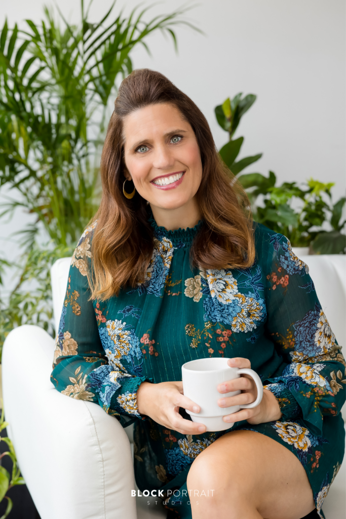Lively portrait of a Caucasian woman with brown hair, wearing a blue floral shirt, sitting on a white couch with greenery in the background, smiling at the camera, and holding a mug, taken by Minneapolos-St. Paul professional photographer Block Portrait Studios.