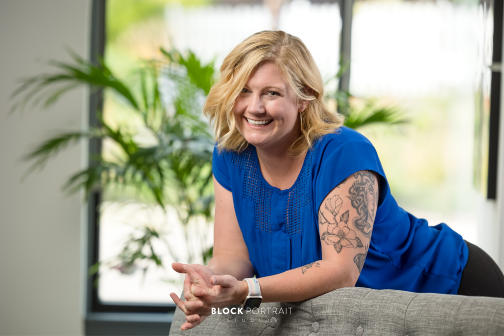 Professional portrait of a blonde, Caucasian woman, with tattoos on her arm, wearing a blue shirt and leaning over a couch smiling by Twin Cities Photographer.