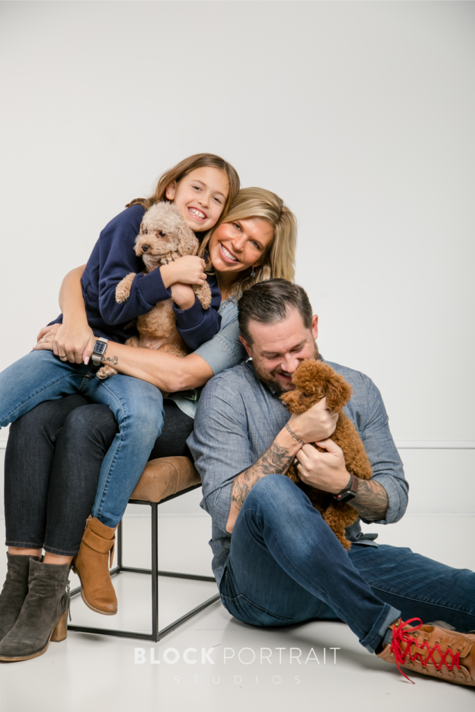 Family portrait of Caucasian family of a wife, husband and young daughter, sitting closely in a white room with their two pet puppies smiling and laughing.