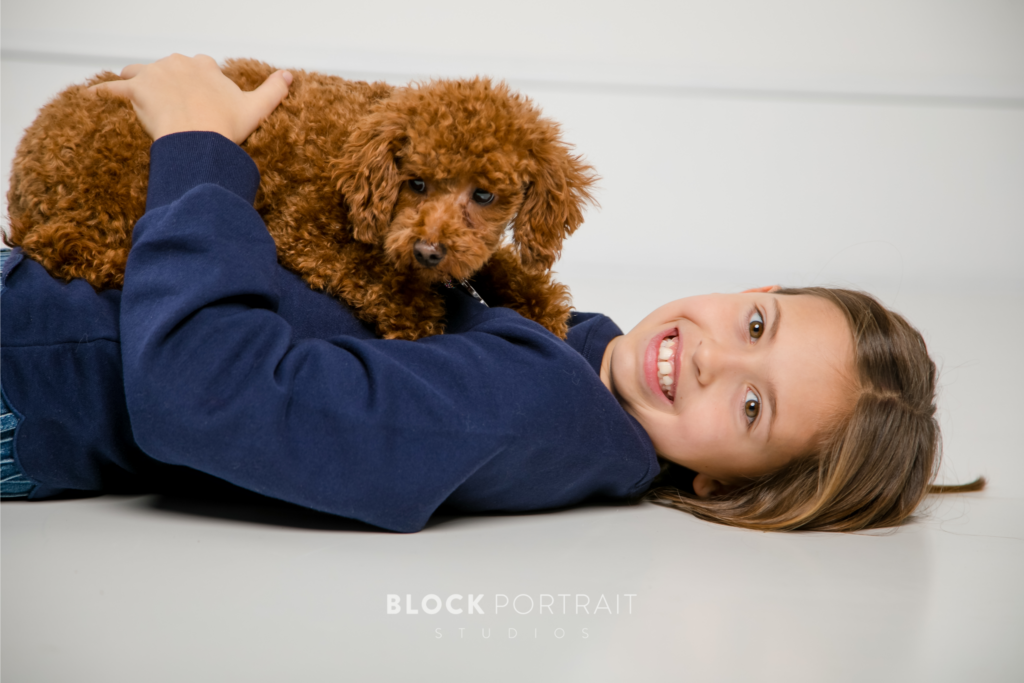 Caucasian girl lying on the floor wearing a navy blue sweater, with her rust brown teddy bear pet dog lying on top of her while she is smiling.