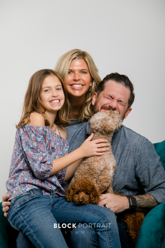 Photo of a Caucasian husband and wife and their child daughter smiling in a white room with one of their two small breed dogs licking the husand's face by St. Paul pet portrait photographer.