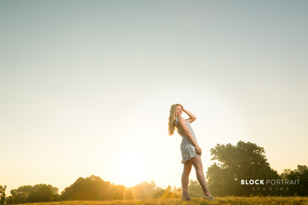 Photo of a blonde high school senior girl posing outside with the sunset and trees behind her, wearing a white and blue floral dress by St. Paul Photographer.