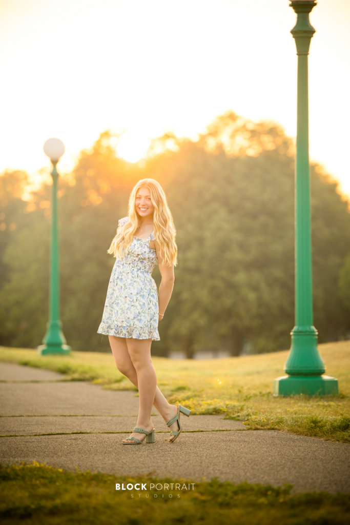 Photo of a Caucasian blonde girl posing outside with the sunset, trees and green light posts behind her, wearing a white and blue floral dress by St. Paul senior portrait Photographer.