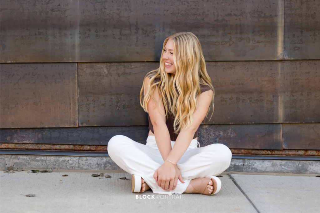 portrait of Caucasian girl with blonde, wavy hair, wearing white jeans and a t-shirt, sitting cross-legged on a sidewalk in front of a brick wall, smiling away from the camera, taken by St. Paul photography studio Block Portrait Studios.