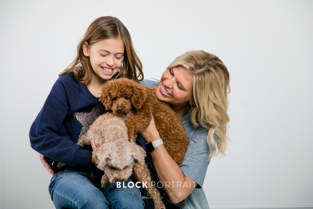 Portrait of blonde Caucasian mother and young daughter, smiling at their two teddy bear puppies in a white room.