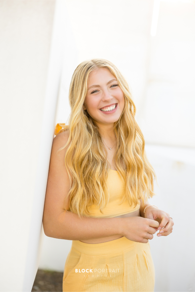 Senior picture of a Caucasian girl with long wavy blonde hair, wearing a set two piece outfit, smiling and posing against a white wall.