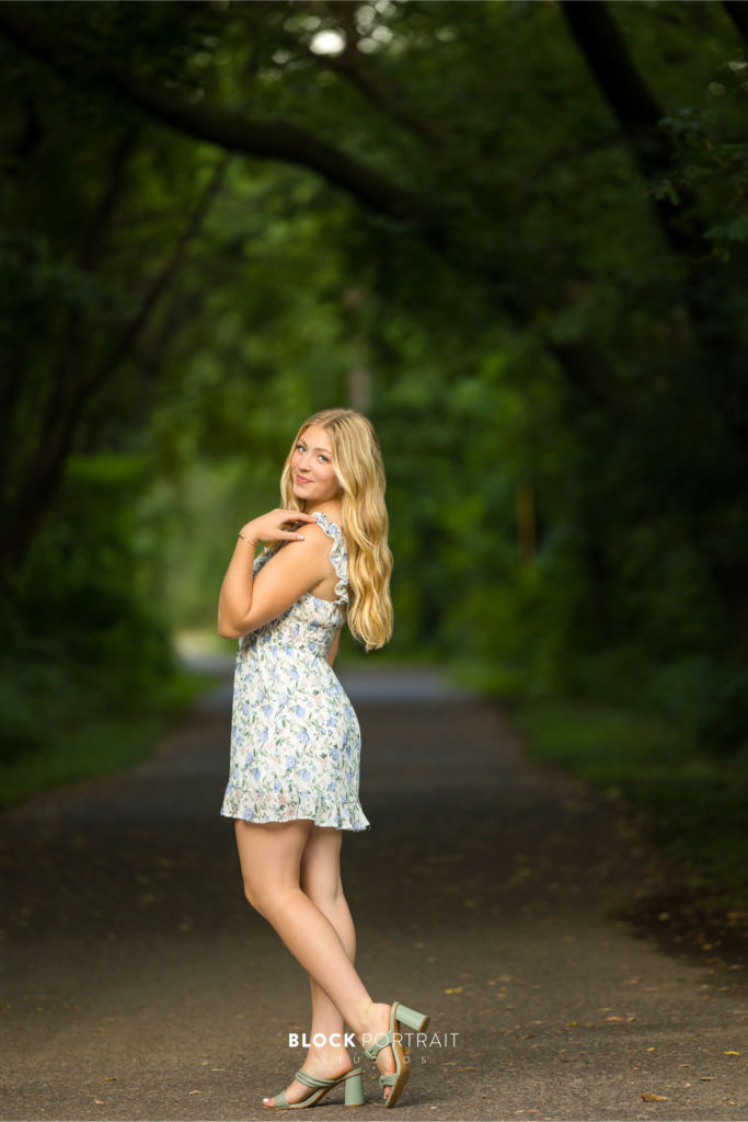 Photo of a Caucasian blonde girl posing outside in the middle of a tree covered street, wearing a white and blue floral dress by Twin Cities senior picture photographer.