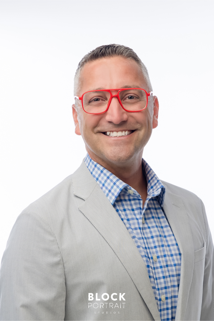 One of a few men's business headshots of a Caucasian man, smiling in front of a white background, wearing bright red framed glasses, and a beige jacket over a blue, plaid shirt taken by Twin Cities photography studio Block Portrait Studios.
