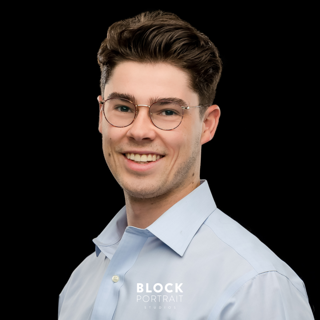 Photo of a Caucasian male posing for his men's business headshots wearing glasses, and a blue collared, button up shirt, standing and smiling in front of a black background taken by Saint Paul portrait photographer, Block Portrait Studios.