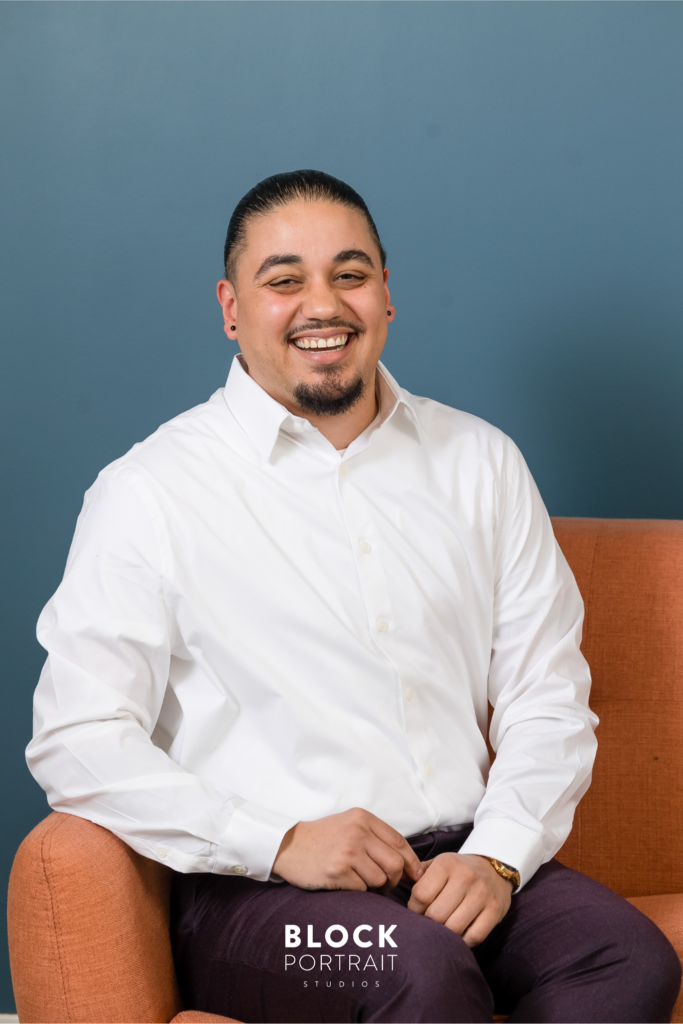 Photo of a Hispanic male posing for his men's business headshots with slicked back black hair, and a mustache and beard, wearing a white button up shirt, sitting on an orange couch with a blue wall.