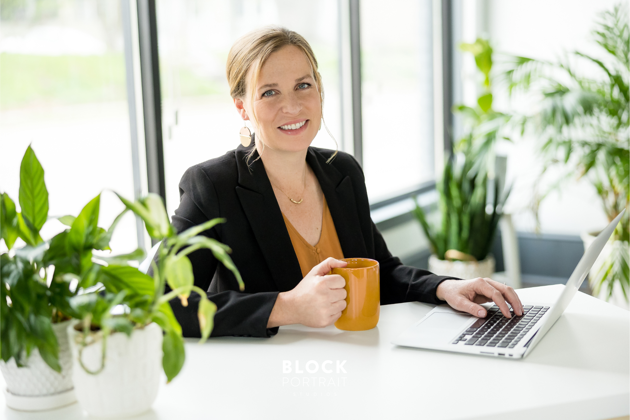 Professional portrait of caucasian woman with blonde hair and blue eyes wearing a black blazer and yellow blouse holding a yellow mug, sitting at a desk with windows behind her and two plants in the background, smiling at the camera, taken for business branding at Block Portrait Studios in Saint Paul, MN.