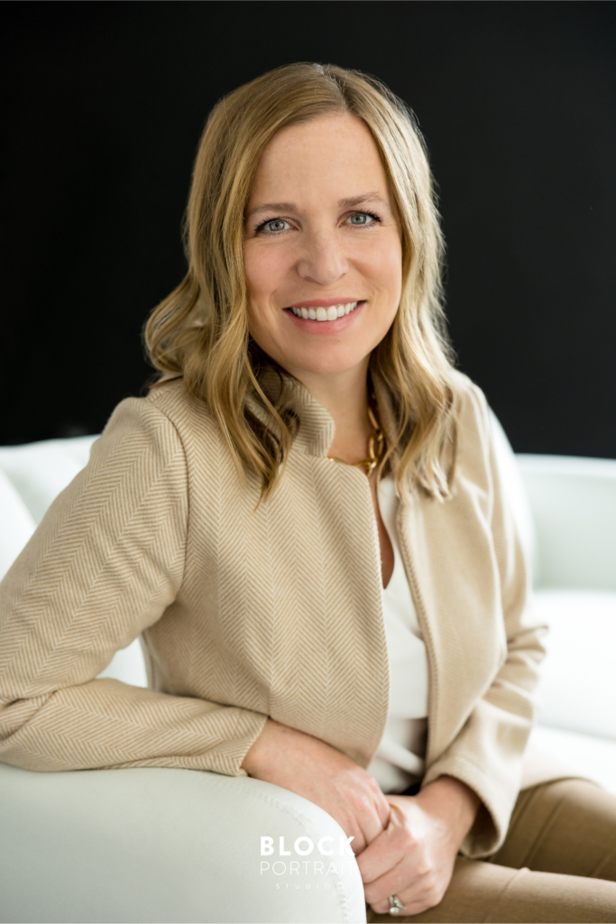 Headshot of a caucasian woman with blonde hair and blue eyes wearing a beige blazer and a white top, sitting on a white couch, smiling at the camera by Block Portrait Studios.