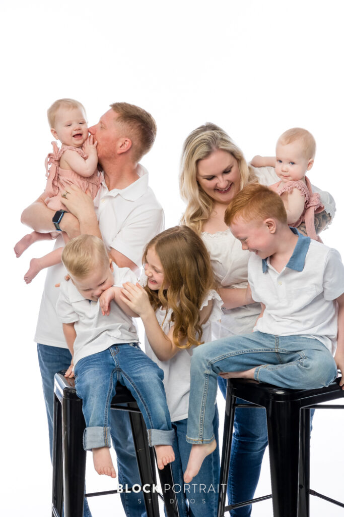 Family portrait taken by Block Portrait Studios in Saint Paul, MN, of a Caucasian family of six; two adult parents and four children with blonde and red hair all under the age of seven, all wearing white shirts and blue jeans. The family is posed in front of a white backdrop, with two of the children sitting on black stools and the rest are positioned around, while they are all joking around and laughing with one another, captured by Block Portrait Studios, a family photography studio in Saint Paul.