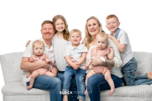 Family portrait taken by Block Portrait Studios in Saint Paul, MN, of a Caucasian family of six; two adult parents and four children with blonde and red hair all under the age of seven, all sitting in front of white backdrop on a grey couch, smiling with the children sitting on the parent's laps, all wearing white shirts and blue jeans.