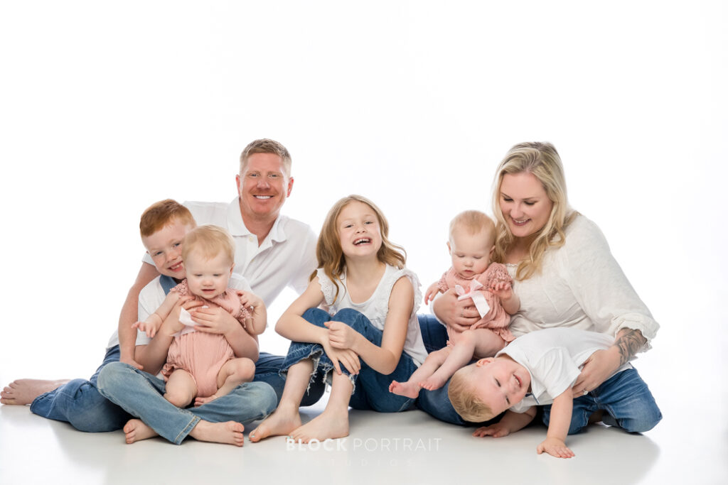 Family portrait taken by Block Portrait Studios in Saint Paul, MN, of a Caucasian family of six; two adult parents and four children with blonde and red hair all under the age of seven, sitting and laughing in front of plain white backdrop, wearing white shirts and blue jeans.