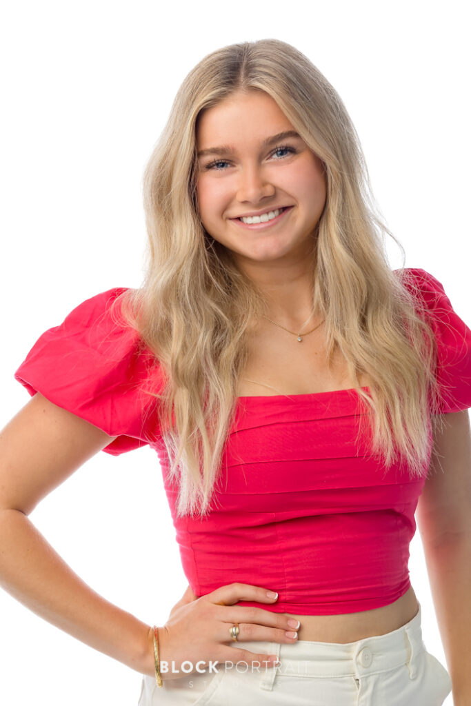 Senior portrait of a Caucasian girl with blonde hair, wearing a coral pink shirt with white jeans, standing, posing, and smiling in front of a white backdrop.