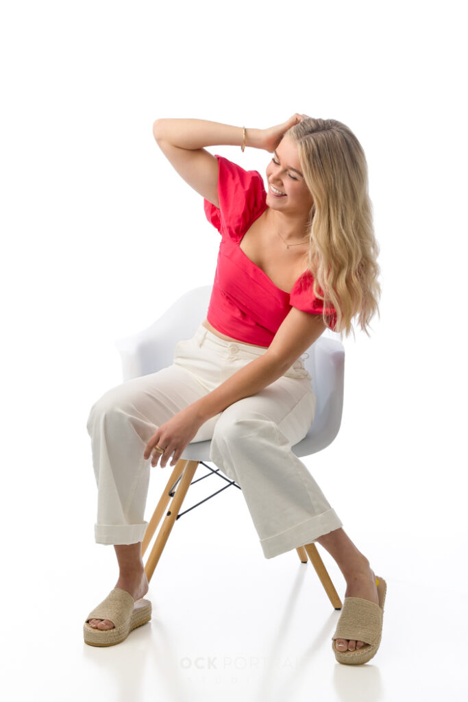 Senior portrait of a Caucasian girl with blonde hair, wearing a coral pink shirt with white jeans, sitting on a white chair in front of a white backdrop, looking away from the camera and running her fingers through her hair, captured by Block Portrait Studios, senior portrait studio in Saint Paul Minnesota, during their Mother's Day Giveaway Session.