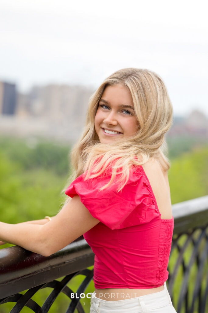 Senior portrait during a family portrait session, of a Caucasian teenage girl with blonde, wavy hair, standing against a railing outside with trees in the background, wearing a coral pink top, looking over her shoulder and smiling at the camera, taken by Block Portrait Studios in Saint Paul, MN.