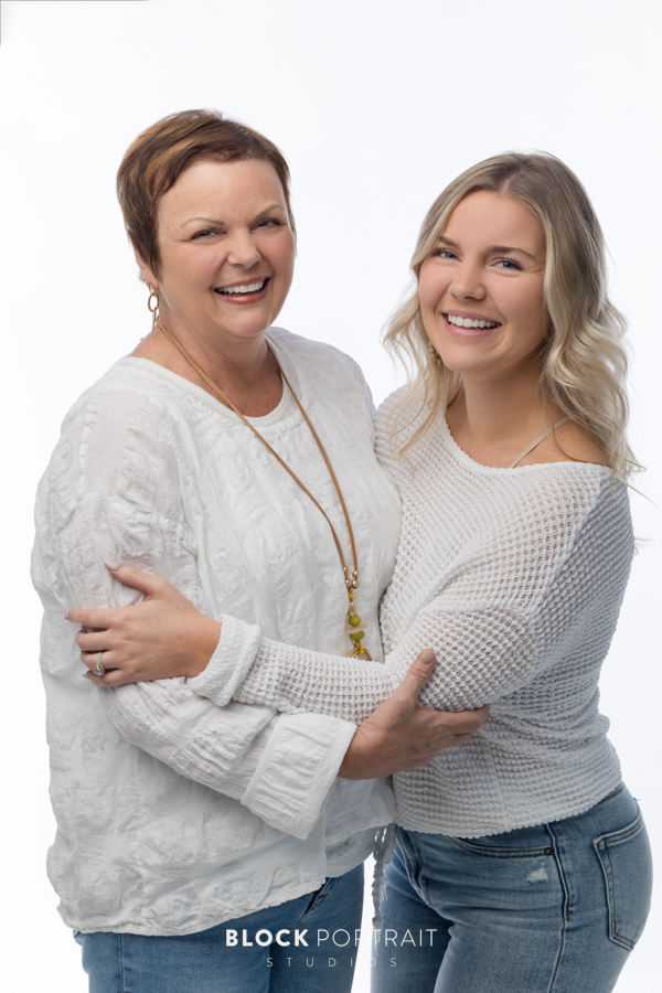 Family portrait of a Caucasian mother with short brown hair, and her Caucasian daughter with curly blonde hair, posing, hugging and smiling in front of a white backdrop, both wearing white long sleeve shirts, and blue denim jeans, taken by family photography studio, Block Portrait Studios, in the Twin Cities in Minnesota.