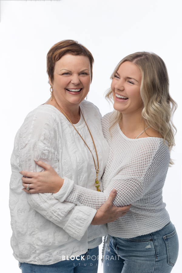 Family photo of a Caucasian mother with short brown hair, and her Caucasian daughter with curly blonde hair, posing, hugging and laughing in front of a white backdrop, both wearing white long sleeve shirts, and blue denim jeans, taken by family photography studio, Block Portrait Studios, in the Twin Cities in Minnesota.