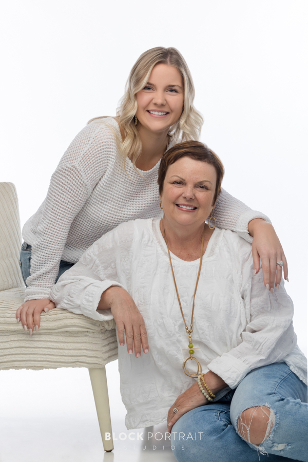 Family photo of a Caucasian mother with short brown hair, and her Caucasian daughter with curly blonde hair, both wearing long sleeve white shirts with blue jeans. The daughter is sitting on a white chair and the mother is sitting on the floor, in front of a white backdrop, posing with their arms around each other and smiling at the camera, captured by Block Portrait Studios in Saint Paul, MN.