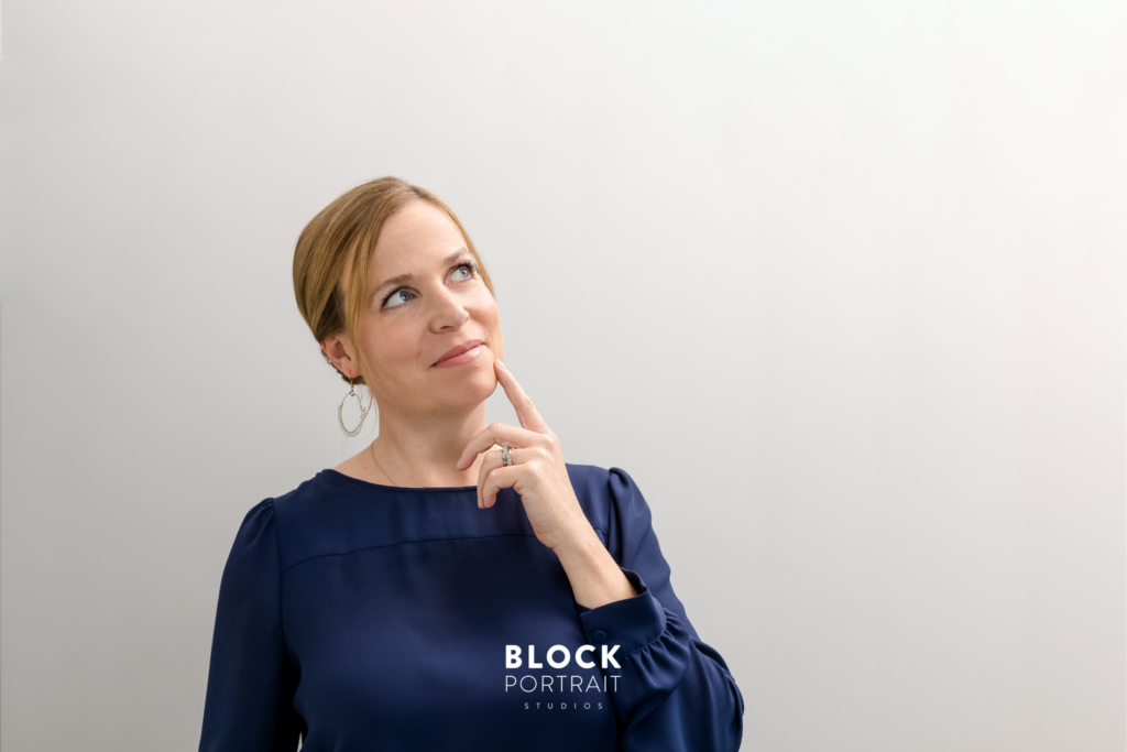 Professional headshot for business branding of a caucasian woman with blonde hair and blue eyes, wearing a navy blue blouse, posing as if she is thinking in front of a white wall taken by Block Portrait Studios.