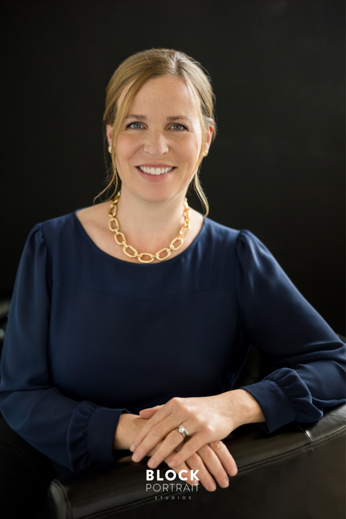 Professional headshot of a caucasian woman with blonde hair and blue eyes, wearing a navy blue blouse and a chunky gold link chain necklace, sitting on a black leather couch with a black backdrop, smiling at the camera, taken by professional photography studio Block Portrait Studios, in Minnesota.