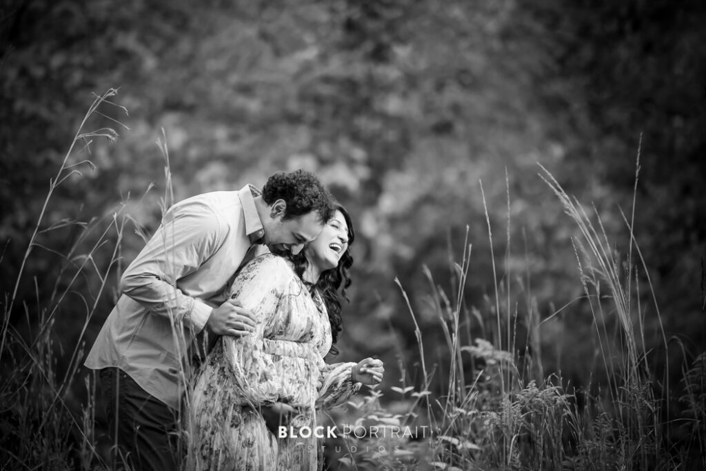 During a maternity photoshoot in St. Paul, MN, the expectant father lovingly back-hugs the mother, both sharing laughter and joy in a tender moment, surrounded by scenic beauty